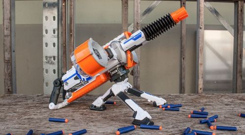 A toy blaster with an orange and white design, positioned on a wooden surface, surrounded by blue foam darts.
