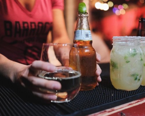 A person holds a glass of clear liquid and a beer bottle, with a cocktail in a jar on the bar.