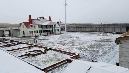 Historic building surrounded by a snowy landscape, with wooden structures and a tall flagpole in a fenced area.
