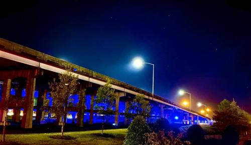 Nighttime view of a highway overpass illuminated by blue and orange lights, with trees and stars in the background.