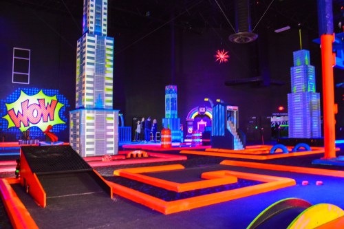A vibrant indoor mini-golf course with neon colors, featuring tall cityscape structures and playful obstacles.