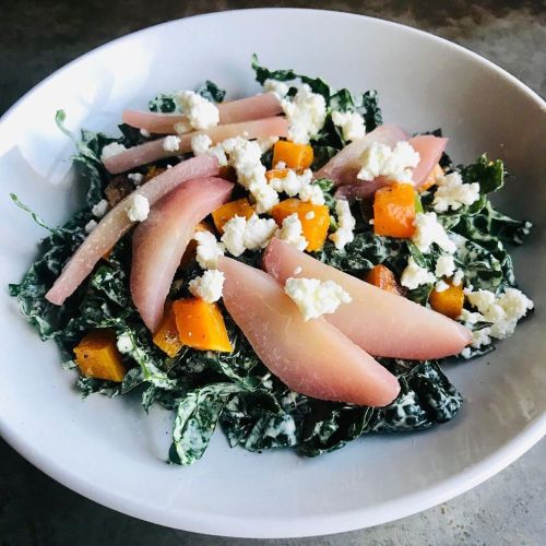 A bowl of kale salad topped with roasted butternut squash, pickled pears, and crumbled feta cheese.