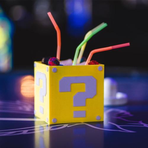 A colorful drink served in a yellow question mark box with multiple straws, set on a vibrant table.