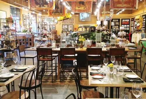 A vibrant restaurant interior featuring a vintage car, art, and elegantly set dining tables with glassware and flowers.