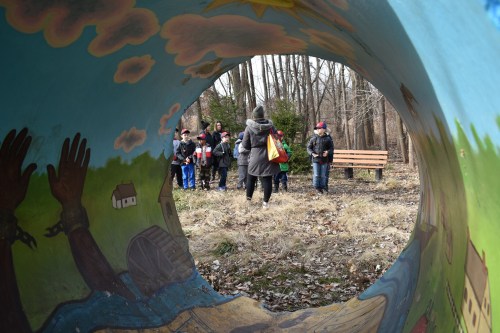 A group of children and adults gather in a park, viewed through a colorful painted tunnel. Trees and a bench are in the background.