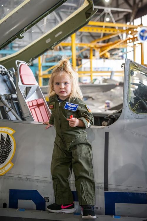 A young child in a flight suit stands by an open cockpit of an aircraft, holding a small toy.