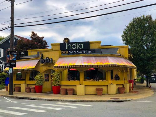 A vibrant yellow restaurant named "India" with outdoor seating and colorful awnings, set against a cloudy sky.