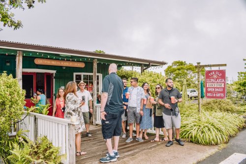 A group of people gathered outside a green building with a sign for a deli and gifts, enjoying a sunny day.