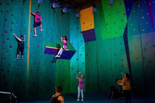 Three climbers ascend a colorful indoor rock wall, while others observe and assist below.