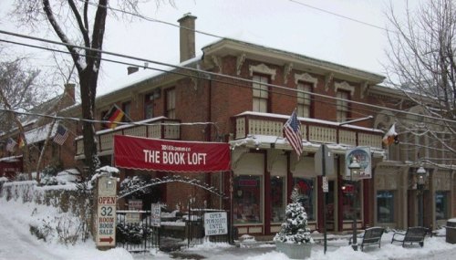 A cozy bookstore, "The Book Loft," covered in snow, with flags and a festive sign, set in a charming winter scene.