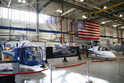 Two helicopters on display in a hangar, with a large American flag in the background and a "Making History" banner.