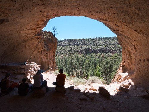 View from inside a cave, showing people sitting and a landscape of trees and hills outside in bright sunlight.
