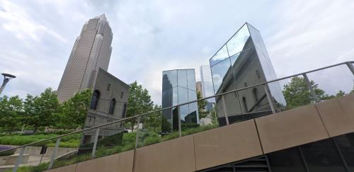 Modern glass buildings reflect the skyline, surrounded by greenery and a sloped walkway in an urban setting.