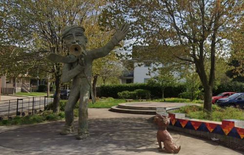 A large statue of a man with an outstretched arm stands in a park, surrounded by trees and a small dog sculpture nearby.