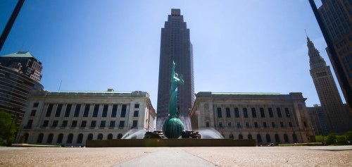 A green statue stands in a plaza, framed by tall buildings under a clear blue sky.