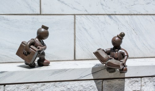 Two bronze sculptures of stylized figures: one holding a briefcase and the other reading a book, set against a marble wall.