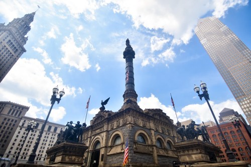 A tall monument with statues, surrounded by buildings and flags, under a blue sky with clouds.