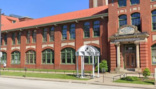 Historic brick building with large windows and a sign reading "Woolfex Company" at the entrance. Green lawn in front.