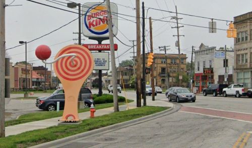 A colorful lollipop sculpture stands at an intersection near a Burger King and various shops on a cloudy day.