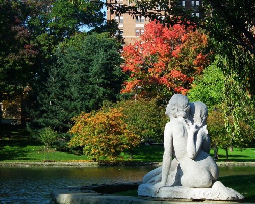 A stone sculpture of two figures sitting back to back, surrounded by trees and a serene water view.