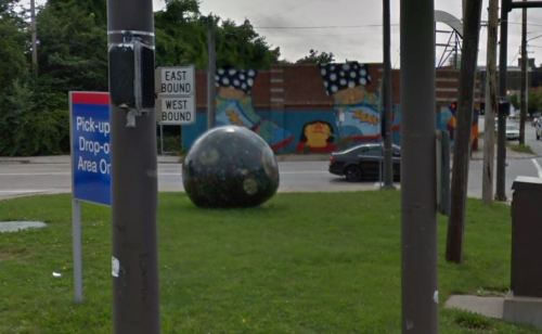 A large, dark spherical sculpture on grass near a street sign and a colorful mural in the background.