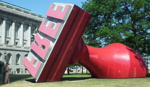 A large red sculpture of a stamp with the word "FREE" tilted on its side in a grassy area near a building.