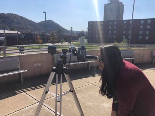 A person looks through a telescope on a sunny day, with buildings and mountains in the background.