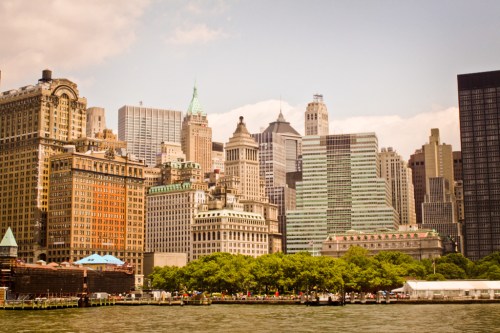 A skyline view of tall buildings in a city, with greenery in the foreground and a clear sky above.