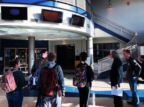 A group of students with backpacks gathers in a school lobby, looking at screens displaying information.