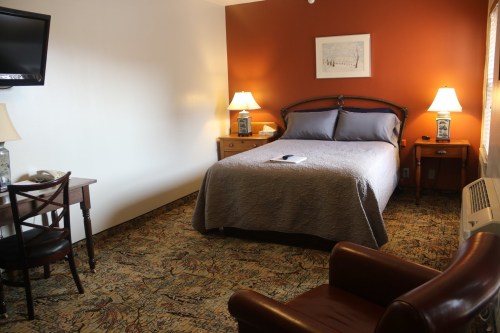 Cozy hotel room with a queen bed, desk, armchair, and lamps, featuring warm orange walls and patterned carpet.