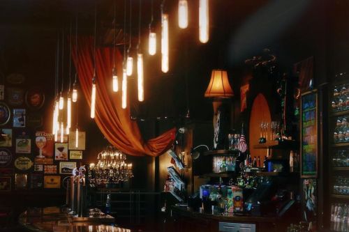 A dimly lit bar with hanging light bulbs, colorful drink displays, and decorative elements on the walls.