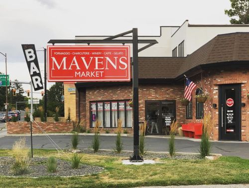 A red sign for Mavens Market, featuring a bar sign, with a brick building and decorative plants in the foreground.