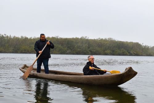 Two men paddle a wooden canoe on a calm river surrounded by trees under a cloudy sky.