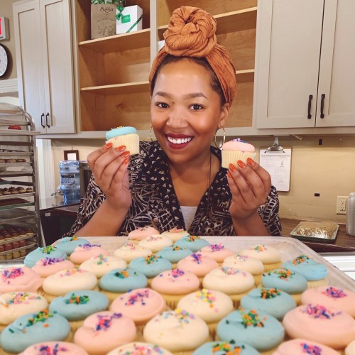 A smiling woman holds two cupcakes in a bakery, surrounded by a tray of colorful frosted cupcakes.