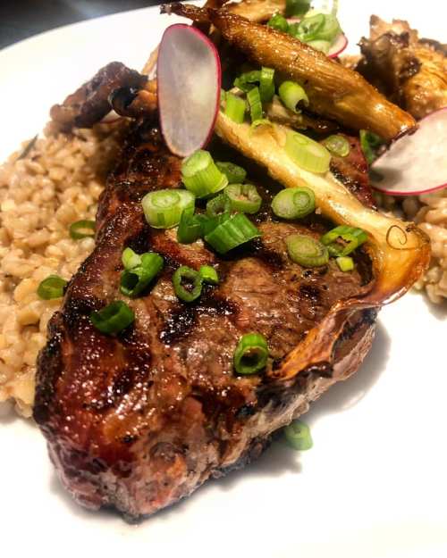 A close-up of grilled steak on a bed of grain, topped with green onions and radish slices, with roasted mushrooms.