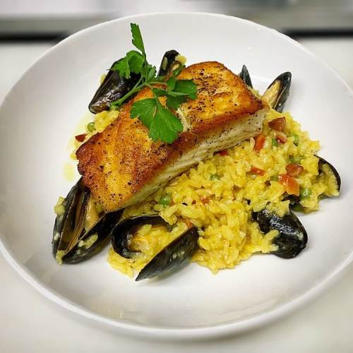 A plate of golden risotto topped with a crispy fish fillet and mussels, garnished with a sprig of parsley.