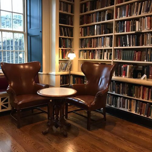 Cozy reading nook with two leather chairs and a small table, surrounded by shelves of books. Warm, inviting atmosphere.