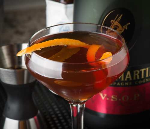 A close-up of a cocktail in a glass, garnished with orange peel, next to a bottle of Martell V.S.O.P. cognac.