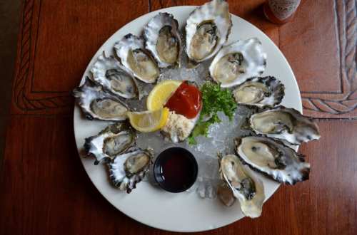 A plate of fresh oysters on ice, garnished with lemon, parsley, and a small dish of sauce.
