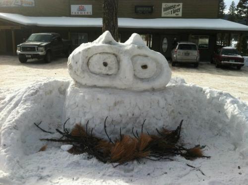 A large snow sculpture resembling a creature with big eyes, surrounded by pine branches, set in a snowy landscape.