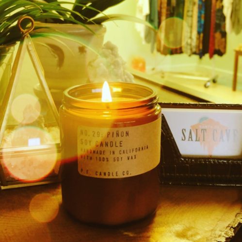 A lit soy candle in a glass jar, labeled "No. 29: Piñon," with a cozy background featuring a plant and a sign.