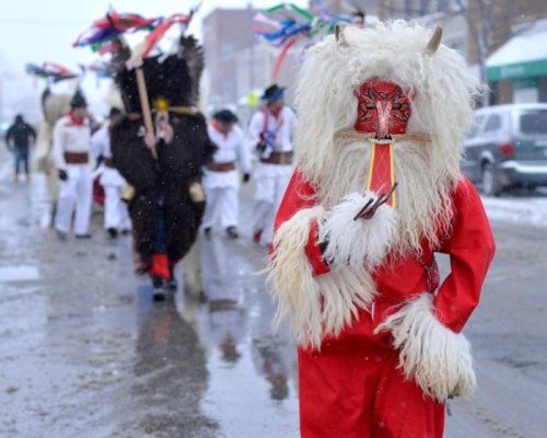 A costumed figure in a red outfit and mask leads a snowy parade, with others in traditional attire following behind.