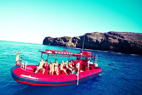 A red boat with several people enjoying the sun on clear blue water near rocky cliffs.