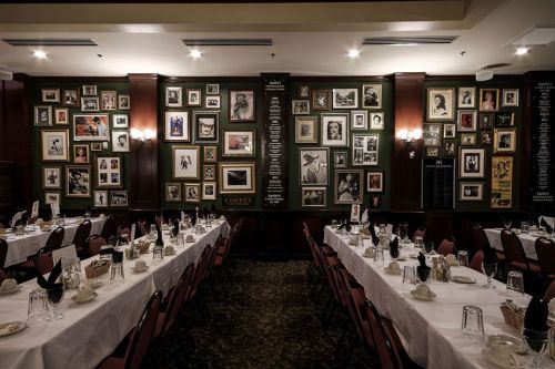 A restaurant interior featuring long tables set for dining, surrounded by a wall of framed black-and-white photos.