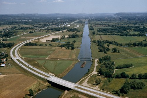 Aerial view of a winding river bordered by fields and a highway, with a bridge crossing over the water.