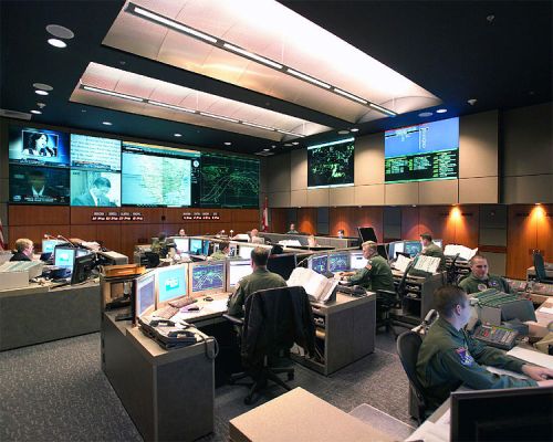 A busy command center with personnel at computers, large screens displaying data and maps, and a high-tech environment.