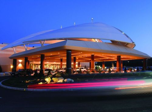 Modern building entrance with a large, curved canopy, illuminated at dusk, surrounded by parked cars and landscaping.
