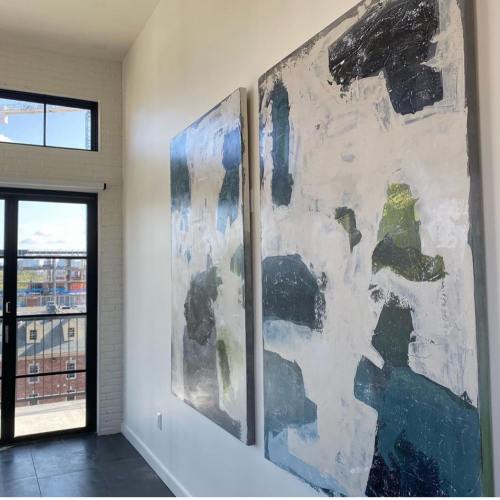 Two large abstract paintings hang on a white wall, with a view of a cityscape through a nearby window.