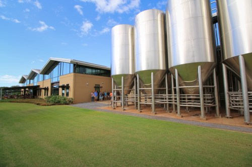 A modern building with large silver silos, surrounded by green grass and a clear blue sky. People are gathered outside.