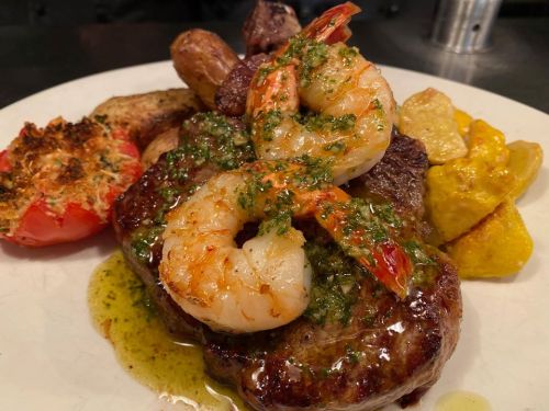 A plate of grilled steak and shrimp topped with green sauce, accompanied by roasted potatoes and a stuffed tomato.